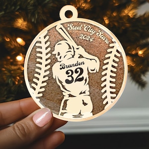 May include: A gold baseball-shaped ornament featuring a silhouette of a baseball player swinging a bat. The ornament is inscribed with the text "Steel City Stars 2024" and "Brandon 32".