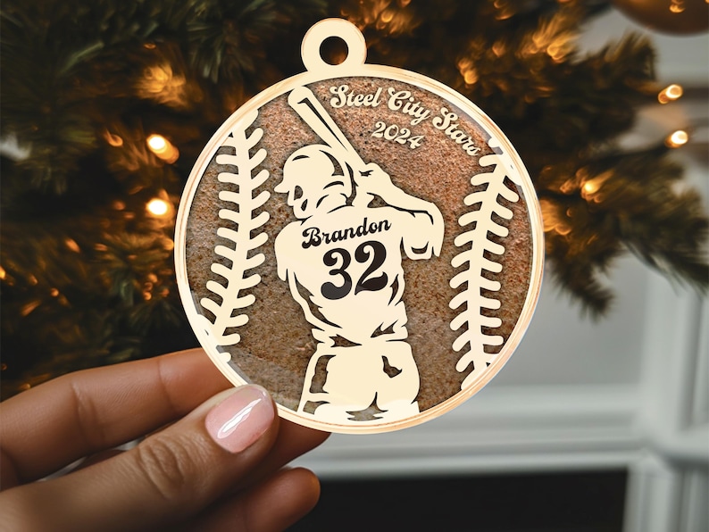 May include: A gold baseball-shaped ornament featuring a silhouette of a baseball player swinging a bat. The ornament is inscribed with the text "Steel City Stars 2024" and "Brandon 32".