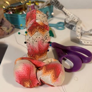 May include: A handmade pincushion with a colorful patchwork design in shades of red, orange, and cream. The pincushion is filled with pins of various colors and sizes. A pair of purple scissors and a measuring tape are also visible.