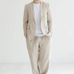 Puede incluir: Un traje de lino beige claro, compuesto por una americana y un pantalón a juego, combinado con una camiseta blanca y zapatillas rojas. El traje tiene un cierre clásico de dos botones y un corte relajado. Adecuado para una ocasión informal o semiformal.