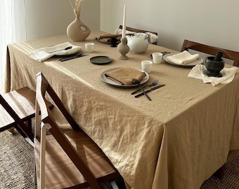 May include: A beige linen tablecloth with a simple, minimalist table setting. The table is set with white plates, black cutlery, and a small brown ceramic vase with dried flowers. There is a teapot and a small mortar and pestle on the table.
