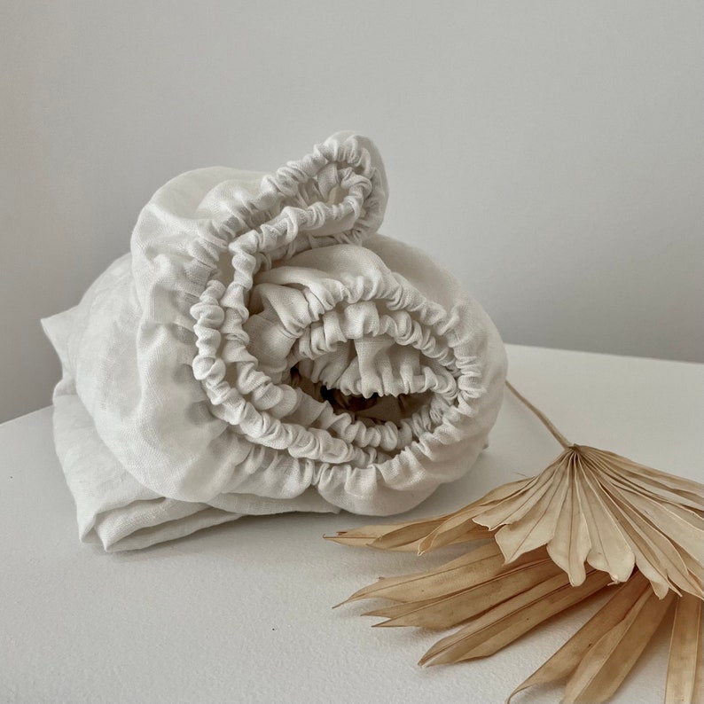 May include: A white linen fitted sheet is rolled up and displayed on a white surface with a dried palm leaf next to it.