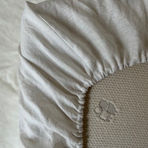 May include: Close-up of a white linen fitted sheet with elastic edging. The sheet is made of a soft, natural fabric and is perfect for a comfortable night's sleep.