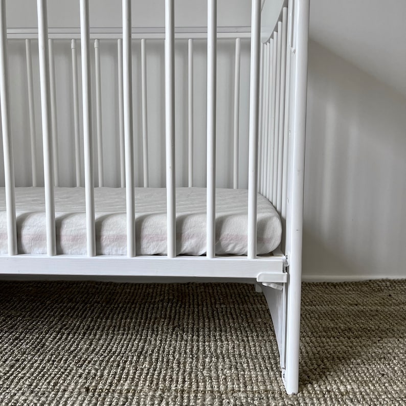 May include: White wooden crib with a white and pink striped mattress. The crib is on a light brown carpet.
