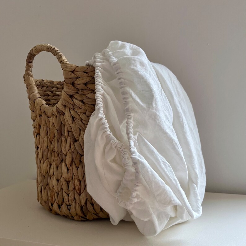 May include: A white linen basket liner in a woven wicker basket. The liner is gathered at the top and has a ruffled edge.