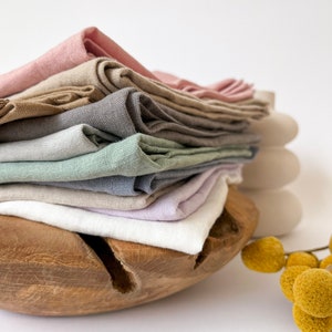 Peut inclure: Une pile de serviettes en lin pliées de différentes couleurs pastel, dont rose, beige, gris et bleu. Les serviettes sont disposées dans un bol en bois avec une finition naturelle. Une branche de fleurs jaunes et des œufs décoratifs blancs sont en arrière-plan.
