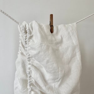 May include: White linen fitted sheet hanging on a clothesline with a wooden clothespin.