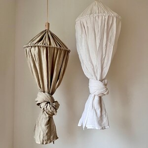 May include: Two hanging fabric canopies, one in a light brown color and the other in white. Both canopies are made of a soft, flowing fabric and have a gathered top with a wooden ring.