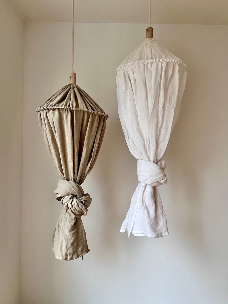 May include: Two hanging fabric canopies, one in a light brown color and the other in white. Both canopies are made of a soft, flowing fabric and have a gathered top with a wooden ring.
