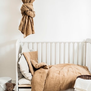 May include: A white crib with a brown linen duvet and pillows. A brown teddy bear sits on a white stool next to the crib. A brown wicker basket with white towels sits on the floor next to the crib.