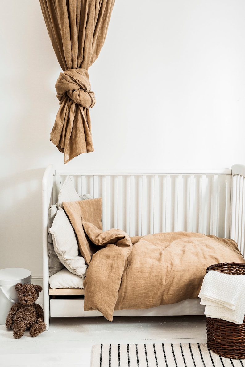 May include: A white crib with a brown linen duvet and pillows. A brown teddy bear sits on a white stool next to the crib. A brown wicker basket with white towels sits on the floor next to the crib.