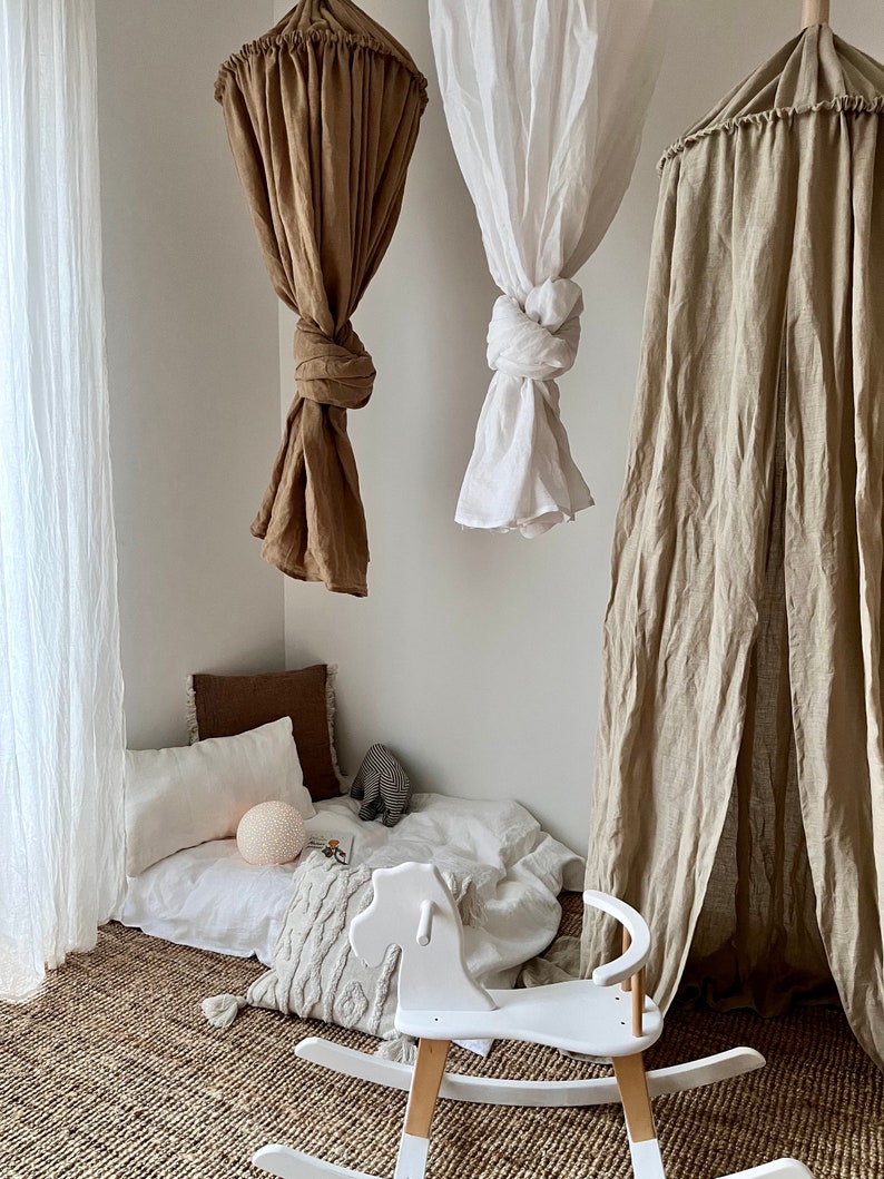 May include: A white rocking horse with wooden rockers sits on a woven rug in a playroom. Three linen canopy tents hang from the ceiling, one white, one brown, and one beige.