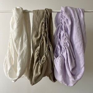 May include: Three fitted bed sheets hanging on a clothesline. The sheets are made of linen and are in different colors: white, beige, and lavender.