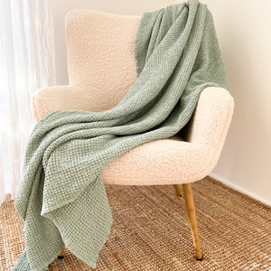 Peut inclure: Une couverture en tissu gaufré vert sauge drapé sur un fauteuil crème avec des pieds dorés. Le fauteuil est posé sur un tapis en fibres naturelles.