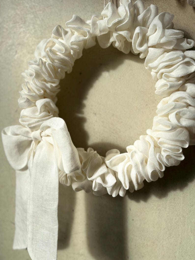 Puede incluir: Una corona de tela blanca con un gran lazo blanco. La corona est&aacute; hecha de tela fruncida y es perfecta para un estilo de decoraci&oacute;n r&uacute;stico o de granja.