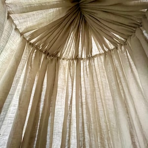 May include: A white sheer fabric canopy with a gathered top, creating a soft and airy look. The fabric is draped in a circular pattern, creating a sense of privacy and intimacy.