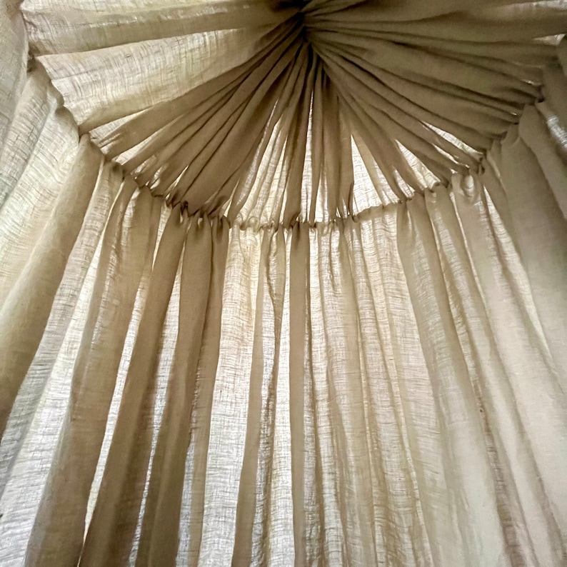 May include: A white sheer fabric canopy with a gathered top, creating a soft and airy look. The fabric is draped in a circular pattern, creating a sense of privacy and intimacy.