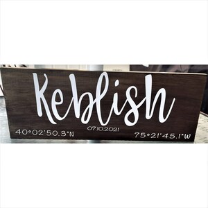 May include: A rectangular, dark stained wooden sign with the name "Keblish" in white cursive lettering. The date "07.10.2021" and coordinates are also displayed in white.