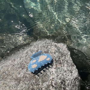 Op de afbeelding: Een lichtblauwe plastic haarspeld met een wolkendesign. De speld ligt op een grijze rots.