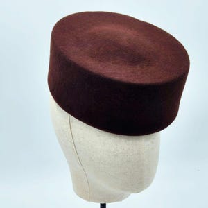 Könnte beinhalten: Ein dunkelburgunderfarbener Hut mit flachem Oberteil, der auf einem Schaufensterkopf ausgestellt ist. Der Hut hat eine zylindrische Form und eine glatte Textur. Der Schaufensterkopf ist hellbeige und der Ständer ist schwarz.