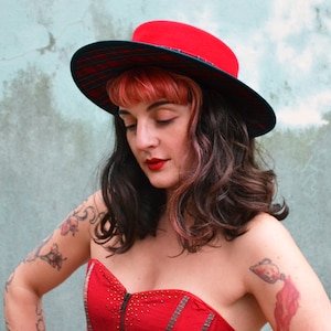 Puede incluir: Una mujer con un sombrero rojo y negro con una banda de cuadros. El sombrero tiene un ala ancha y una corona plana. La mujer lleva un top rojo sin tirantes.