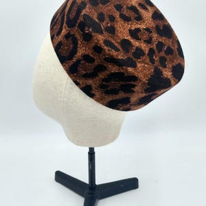 Op de afbeelding: Een bruin en zwart luipaardprint hoed, tentoongesteld op een mannequin hoofd. De hoed heeft een afgeronde kroon en een platte rand. De hoed is gemaakt van een zacht materiaal, waarschijnlijk een modeaccessoire.