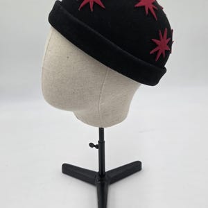 Może przedstawiać: Czarny kapelusz typu cloche z czerwonymi aplikacjami w kształcie gwiazd. Kapelusz ma zaokrągloną koronę i mały, podwinięty rąbek. Kapelusz jest prezentowany na głowie manekina z czarnym metalowym stojakiem.