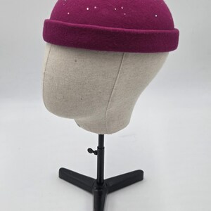 May include: A magenta-colored beanie hat with a rolled brim and scattered silver embellishments. The hat is displayed on a mannequin head, showcasing its shape and design. This hat is a stylish accessory.