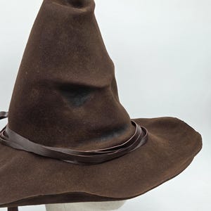 Può includere: Un cappello da mago marrone con tesa larga e corona a punta. Il cappello è adornato da una fascia in pelle marrone attorno alla base della corona. Il cappello è realizzato in un materiale simile al feltro.