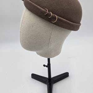Op de afbeelding: Een bruine bowlerhoed met een afgeronde kroon en een smalle rand. De hoed is versierd met drie goudkleurige ringen en een enkele gouden punt. De hoed wordt getoond op een mannequinhoofd.