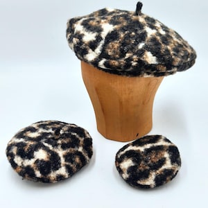 May include: A leopard print beret and two matching round accessories. The beret is displayed on a wooden head form. The pattern features black, brown, and white spots. The accessories are also in the same pattern.