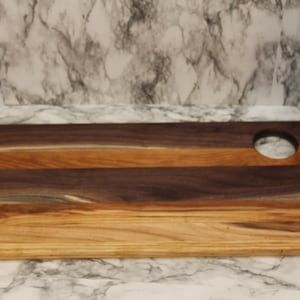 May include: A rectangular wooden cutting board featuring horizontal stripes of dark brown and light tan wood grain. The board has a smooth, flat surface and a circular hole cut out near the top right corner for hanging or easy handling.