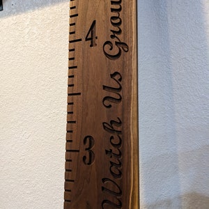 May include: A wooden growth chart with a ruler and the text "Watch Us Grow!" engraved on it. The chart is made of dark brown wood and has a natural wood grain.