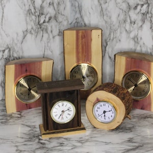 May include: A collection of handcrafted wooden clocks. The clocks feature natural wood grain and bark, with gold-toned clock faces. The wood tones range from light tan to deep brown and reddish-purple. The clocks vary in shape and size, showcasing the natural beauty of the wood.