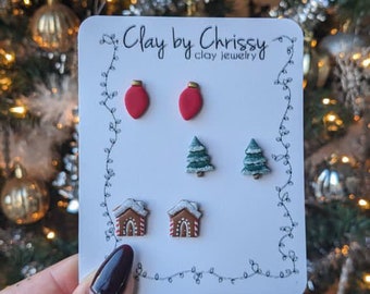 May include: A set of six Christmas-themed clay earrings. The earrings are in the shape of ornaments, Christmas trees, and gingerbread houses. The earrings are displayed on a white card with the text "Clay by Chrissy clay jewelry".