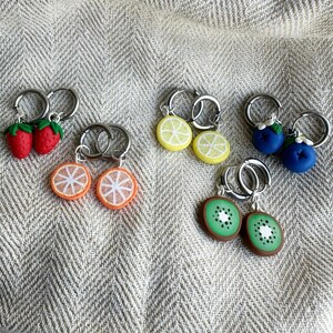 May include: A set of six fruit-shaped earrings. The earrings are made of silver hoops with polymer clay charms. The charms are shaped like strawberries, lemons, oranges, blueberries, and kiwis.