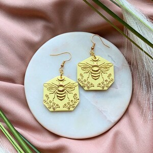 May include: A pair of yellow hexagonal earrings with a gold bee and floral design.