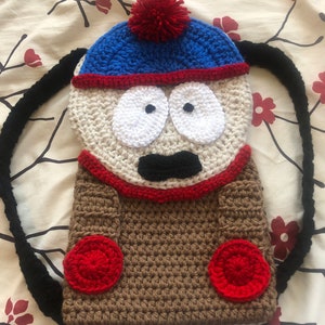 Può includere: Uno zaino all'uncinetto a forma del personaggio Eric Cartman dello show televisivo animato South Park. Lo zaino è marrone con accenti rossi e presenta un viso bianco con occhi neri e una bocca nera. Lo zaino ha un cappello blu con un pon pon rosso in cima.
