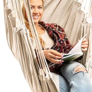 May include: A beige fabric hanging chair featuring a deep seat and integrated side support panels. The chair is suspended by thick, durable white ropes that attach to a central overhead point, creating a structured, comfortable seating area.