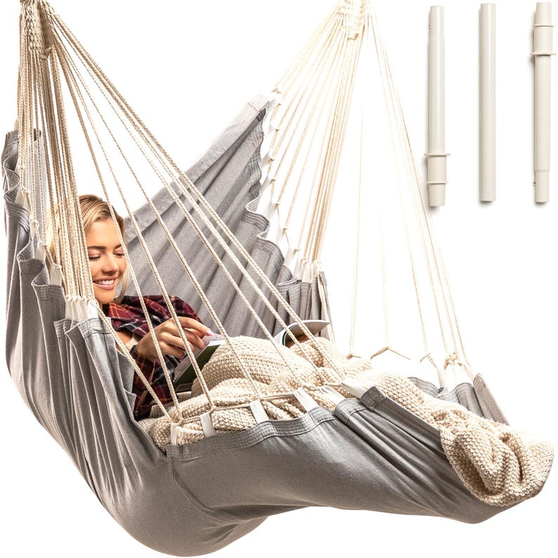 May include: A gray hammock with white ropes and a person sitting in it reading a book. The hammock is hanging from a white stand.
