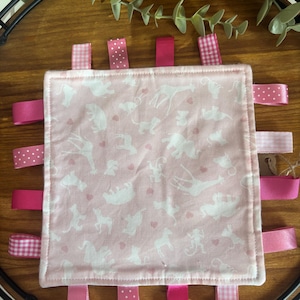 May include: A pink baby tag blanket featuring a white animal pattern and pink ribbons. The square blanket has a white stitched border and various pink ribbons around the edges. Some ribbons are plain pink, others have polka dots or a gingham design.