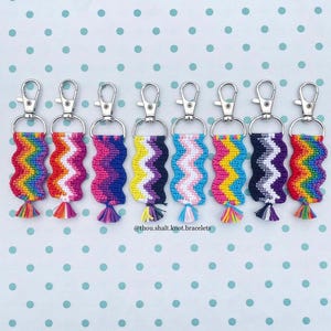Op de afbeelding: Een verzameling handgemaakte sleutelhangers met zilverkleurige sluitingen. Elke sleutelhanger heeft een uniek, kleurrijk, geweven ontwerp in een golvend patroon, met een bijpassende kwast aan de onderkant. De sleutelhangers zijn gerangschikt op een lichtblauwe ondergrond met witte stippen.