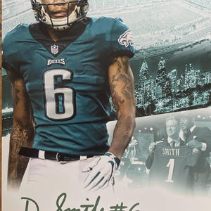 Puede incluir: Impresión enmarcada de un jugador de fútbol americano con uniforme de los Eagles turquesa y blanco, número 6. El jugador lleva casco y guantes. El fondo muestra un estadio y un horizonte urbano. Firmado "D. Smith #6".