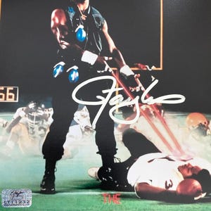 May include: A signed poster featuring a man in a football uniform with the text "The Terminator" in red. The image has a dark background with a football field and other players in the background. The signature is in white.