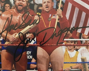 May include: A photograph of two wrestlers in a wrestling ring. One wrestler is holding a wooden object and the other is holding an American flag. Both wrestlers are wearing wrestling attire. The image is signed by the wrestlers.