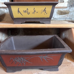 May include: Two rectangular brown ceramic bonsai pots with flared rims and small feet. The top pot features a yellow panel with black Chinese calligraphy. The bottom pot displays a red panel with a black etched bamboo design on the front side.