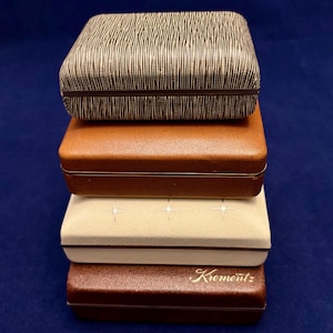 May include: A stack of four vintage jewelry boxes in various colors. The top box has a textured, black and white striped pattern. The other boxes are brown, tan, and dark brown, with the bottom box labeled "Krementz".