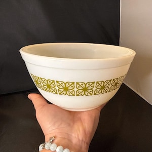 May include: White mixing bowl with a green floral pattern around the exterior. The bowl has a rounded shape and a wide rim. The floral design is a repeating pattern of stylized flowers. The bowl is held in a hand.