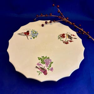 May include: A cream-colored, scalloped-edge serving platter with bird and floral illustrations. The platter features three bird designs with colorful flowers. A sprig of red berries rests on the edge of the platter.