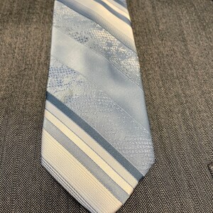 May include: A light blue and white striped tie with various textured patterns. The tie is displayed on a gray herringbone patterned suit. The tie's design includes diagonal stripes and textured sections.